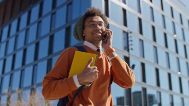 Sırt çantası ve kitaplarla gülümseyen üniversite öğrencisi günışığında modern cam binaların önünde akıllı telefondan konuşuyor..