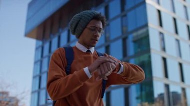 Endişeli genç adam sırt çantası takıyor ve bere dışarıda kol saatine bakıyor, muhtemelen derse ya da randevuya geç kalıyor..