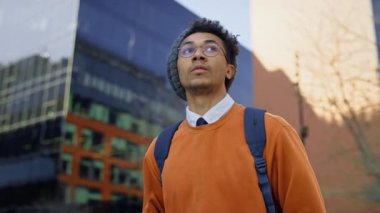 Sırt çantalı genç bir öğrenci modern şehir ortamında yürüyor, yukarı bakıyor, muhtemelen keşfe çıkıyor ya da sınıfa gidiyor..