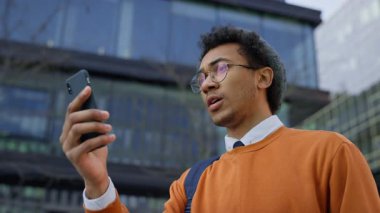 Gözlüklü ve sırt çantalı genç adam telefon ekranına bakıyor, muhtemelen bir video görüşmesinde, modern binaların önünde duruyor..