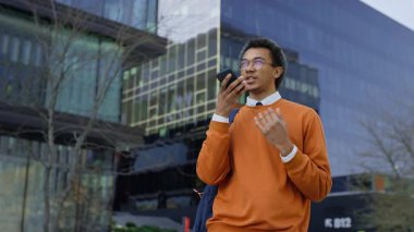 Sırt çantası ve gözlüklü genç adam akıllı telefonu ağzına yakın tutuyor. Modern binaların önünde dikilirken sesli mesaj kaydediyor..