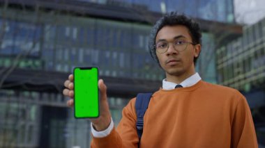 Turuncu tişörtlü genç adam yeşil ekranlı telefonu şehir ortamında duran model veya uygulama ekranı için tutuyor..