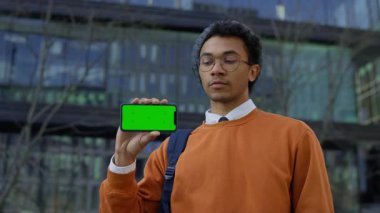 Turuncu kazaklı genç adam telefonu yeşil ekran ile yan tutuyor. Uygulama arayüzünü veya video içeriğini göstermek için ideal..