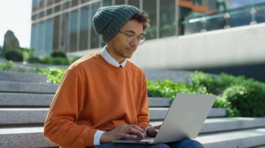 Turuncu sweatshirt ve bereli genç adam kampüs basamaklarında dizüstü bilgisayar kullanıyor, internette geziniyor ya da modern şehir ortamında araştırma yapıyor..