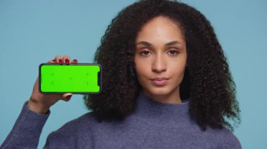 Genç bir kadın yeşil ekran maketli akıllı telefonu tutarken aynı zamanda onaylamayan bir yüz ifadesi takınarak memnuniyetsizlik ya da olumsuz tepki gösteriyor..