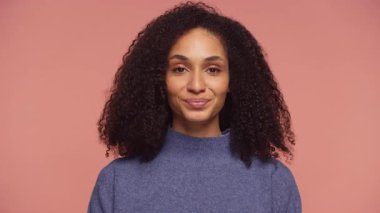 Kıvırcık saçlı, kendinden emin genç bir kadın onaylı bir şekilde başını sallıyor ve gülümsüyor. Yumuşak pembe bir arka plana karşı onay ve pozitifliğini dile getiriyor..