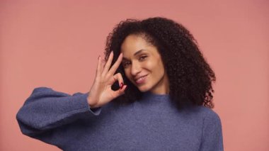 Genç bir kadın kendine güvenerek gülümsüyor ve pembe bir arka plan karşısında onay ya da memnuniyetini belirten bir işaret veriyor..