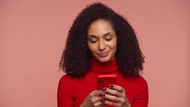 Kırmızı balıkçı yakalı, gülümseyen genç bir kadın. Akıllı telefonuyla mesaj atıyor. Pembe arka plana karşı pozitif tavır ve zevk gösteriyor..