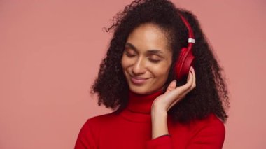 Kırmızı boğazlı genç kadın, gözleri kapalı, pembe arka planda kırmızı kulaklıkla müzik dinlerken huzur içinde gülümsüyor..