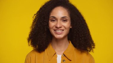 Neşeli, kıvırcık saçlı, kameraya özgüvenle gülümseyen, hardal gömlekli, koyu sarı bir arka planı olan genç bir kadın..