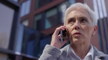 Kısa beyaz saçlı yaşlı kadın modern ofis binalarının dışından ciddi bir telefon görüşmesi yapıyor..