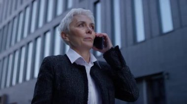 Beyaz saçlı profesyonel yaşlı bir kadın, şehir merkezinde uzun ofis binalarıyla dolaşırken telefon görüşmesi yapıyor..