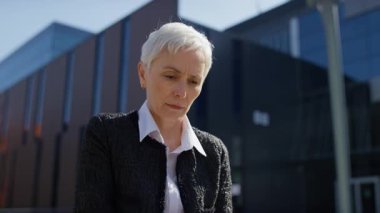 Beyaz saçlı yaşlı kadın, modern bir ofis binasının önünde dizüstü bilgisayar kullanırken stresli görünüyor. İşle ilgili baskıyla karşı karşıya..
