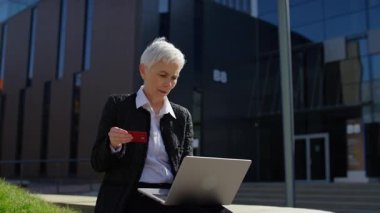 Beyaz saçlı yaşlı kadın kredi kartı bilgilerini girerken gülümsüyor, bir iş bölgesinde online alışveriş veya alım satım yapıyor..