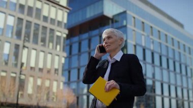 Kendine güveni tam, beyaz saçlı yaşlı bir kadın telefon görüşmesi sırasında gülümsüyor. Elinde sarı bir defter var..