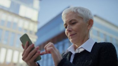 Kentte yürürken akıllı telefon kullanan, sakin ve odaklanmış bir ifadeyle gezinen ya da kaydıran kendinden emin kıdemli iş kadını.