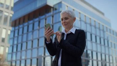 İş kıyafetli neşeli yaşlı kadın dışarıda akıllı telefonunda gördüğü bir şeye şaşırıp seviniyor..