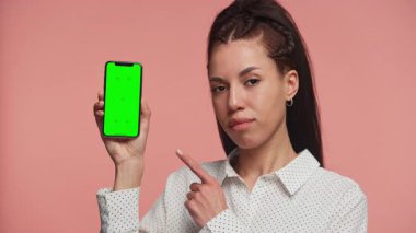 Beyaz bluzlu genç bir kadın yeşil ekranlı bir akıllı telefonu işaret ediyor. Pembe arkaplana karşı uygulama veya ürün yerleştirme öneriyor..