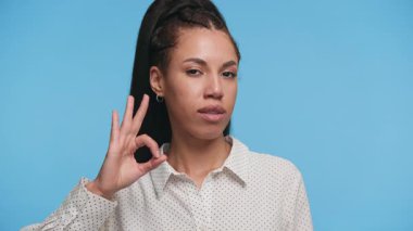 Kendine güveni tam genç bir kadın, mavi arka planda dikilirken, ileri doğru bakarken eliyle 