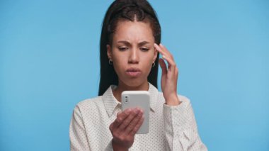 Genç bir kadın akıllı telefonunda mesaj okurken endişeli görünüyor. Stresle tepki veriyor ya da mavi arka planda kafa karışıklığı yaşıyor..