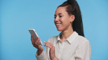 Şaşırmış ve sıkıntılı genç bir kadın elinde akıllı telefon, beklenmedik ve üzücü haberlere tepki verirken mavi arka planda duruyor..