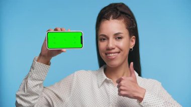 Yeşil ekranlı akıllı telefonu tutarken gülümseyen kadın, memnuniyetini ve pozitif kullanıcı deneyimini ifade ediyor, mavi arka planda izole edilmiş..