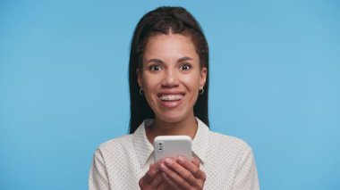 Akıllı telefona bakıp tezahürat eden mutlu genç bir kadın, mavi arka plana rağmen başarıyı ya da iyi haberi kutluyor..