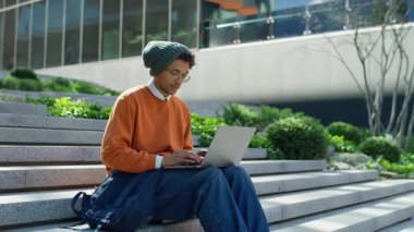 Genç bir öğrenci kampüs merdivenlerinde dizüstü bilgisayar kullanıyor. Gün ışığında çalışıyor ya da ders çalışıyor..