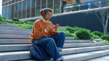 Merdivenlerde oturan, kulaklık takan ve elinde akıllı telefon tutan hayal kırıklığına uğramış erkek öğrenci, olumsuz bir mesaj ya da sınav sonucuna duygusal tepki veriyor..