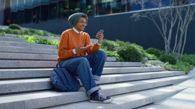 Dışarıda basamaklarda oturan gülümseyen bir öğrenci, elinde akıllı telefon, video görüşmesi sırasında konuşuyor, uzaktan konuşmanın tadını çıkarıyor..
