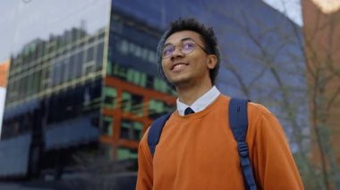 Kendine güvenen bir öğrenci sırt çantasıyla geziyor, gülümsüyor ve etrafındaki uzun camdan binalara bakıyor..