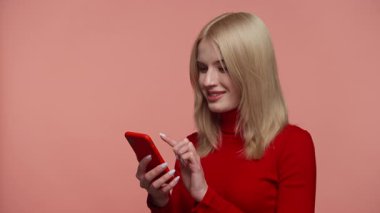 Kırmızı kazaklı genç sarışın kadın elinde akıllı telefon tutarken heyecanlı görünüyor. Pembe stüdyo geçmişine karşı neşeli bir sürpriz yapıyor..