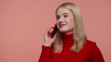 Kırmızı balıkçı yaka yakalı genç sarışın kadın akıllı bir telefonla konuşurken nazikçe gülümsüyor, yumuşak pembe stüdyo arka planına karşı..