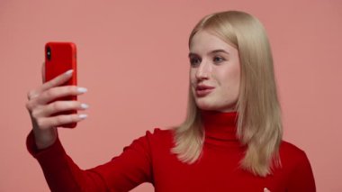 Kırmızı kazaklı sarışın bir kadın önünde akıllı telefon tutuyor, pembe bir arka planla video görüşmesi yaparken konuşuyor..