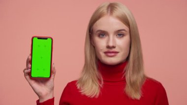 Kırmızı balıkçı yakalı sarışın kadın kameraya doğru yeşil ekranlı akıllı telefonu tutuyor. Pembe arka planda sunum yapıyor..