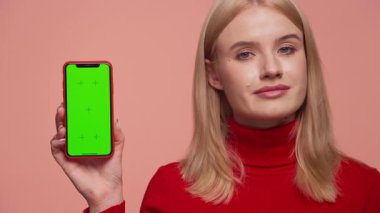 Kırmızı kazaklı sarışın kadın yeşil ekranlı akıllı telefonu tutarken onaylamayarak başını sallıyor. Ürünlerden memnun olmadığını söylüyor. Pembe arka planda izole edilmiş..