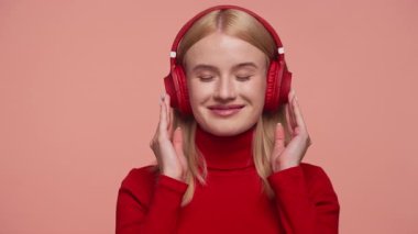 Kırmızı kazaklı sarışın kadın gözlerini kapatır ve kırmızı kulaklıkla müzik dinlerken gülümser. Yumuşak pembe arka planda durur..
