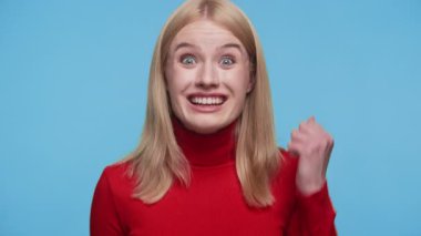 Kırmızı balıkçı yakalı sarışın kadın coşkulu bir ifade sergiliyor ve parlak mavi arka plana karşı yumruğunu sıkıyor..