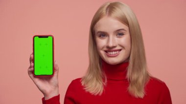 Kırmızı elbiseli sarışın kadın pembe stüdyo arka planında yeşil ekranlı akıllı telefon kullanıyor.