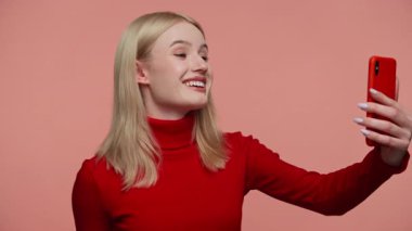 Kırmızı elbiseli sarışın kadın pembe arka planda akıllı telefonuyla konuşurken gülümsüyor.