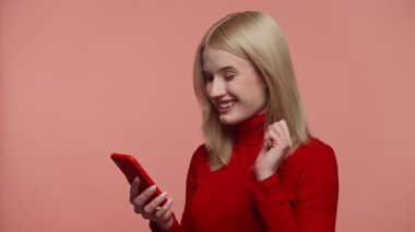 Kırmızı balıkçı yaka kazaklı mutlu sarışın kadın pembe arka planda akıllı telefonu tutarken gülümsüyor ve kutluyor.