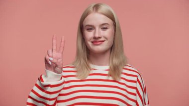 Çizgili tişörtlü sarışın kadın gülümsüyor ve pembe stüdyo geçmişine karşı barış işareti yapıyor.