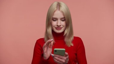 Kırmızı tişörtlü sarışın kadın telefonu pembe arka planda tutarken heyecanla ve neşeyle tepki veriyor.