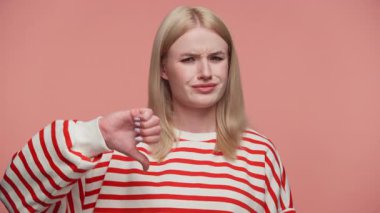 Mutsuz sarışın kadın pembe arka planda kaşlarını çatarak baş parmak hareketi yapıyor.