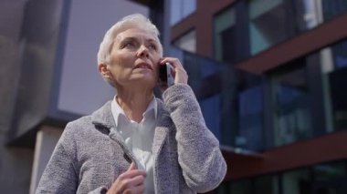 Kısa beyaz saçlı yaşlı kadın akıllı telefondan konuşuyor, ciddi ve endişeli görünüyor, modern ofis binalarının önünde duruyor..