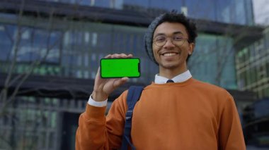 Modern binanın yanında elinde yeşil ekran modeli olan mutlu genç adam kameraya gülümsüyor.