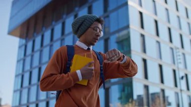 Genç adam aciliyetle ukalalığa bakıyor, elinde defter ve ofis binasının önünde sırt çantası var.