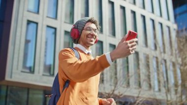 Kulaklıklı mutlu genç adam modern cam binanın dışında akıllı telefondan video görüşmesi yapıyor.