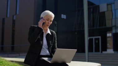 Yaşlı kadın dışarıda dizüstü bilgisayarda çalışıyor ve telefonla konuşuyor, birden fazla iş yapmaya odaklanmış.