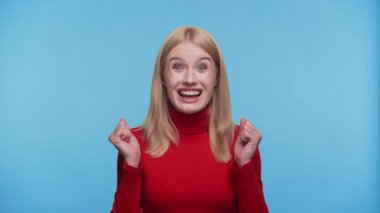 Kırmızı kazaklı sarışın kadın neşeli bir ifadeyle kutlama yapıyor ve mavi arka planda yumruklarını sıkıyor.